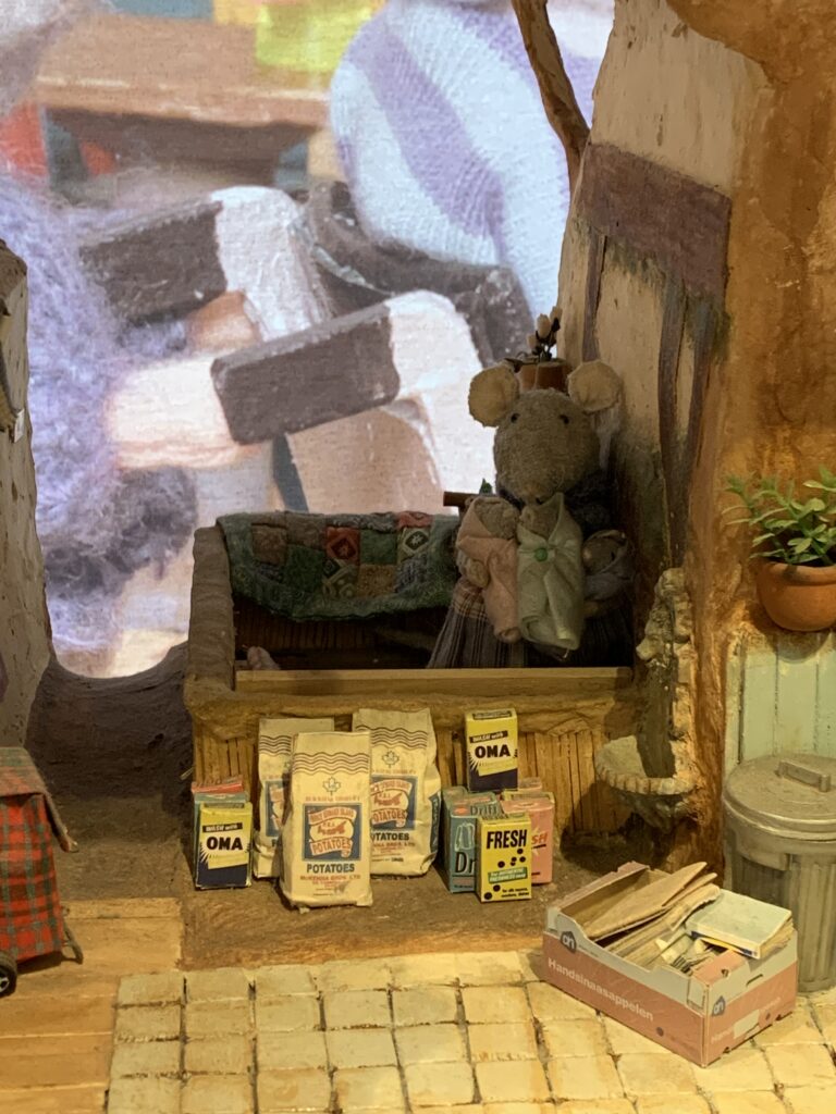 Three small white PEI potato bags are  shown in amongst other tiny replicas of consumer products,  with a tiny mouse holding even tinier baby mice in its hands, in the background, all part of a set from an Amsterdam tourist attraction.