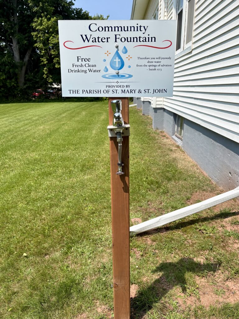 A wooden post that has a water tap attached and a sign that reads: Community Water Fountain. Free Fresh Clean Drinking Water. Therefore you will joyously draw water from the spring of salvation. Isaiah 12:3 Provided by the Parish of St. Mary and St. John.