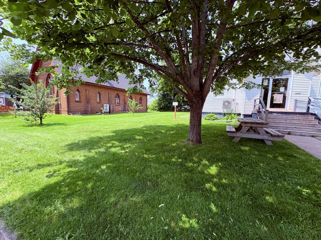 St. Mary’s Anglican Church on the left and the church hall on the right, with a brown picnic table under a tree in front of the hall and green grass.