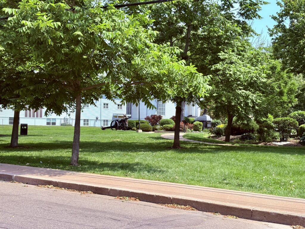 A park with green grass, trees and shrubs, a sidewalk in the foreground, a white church in the background and a person painting a canon in the middle of the photo.