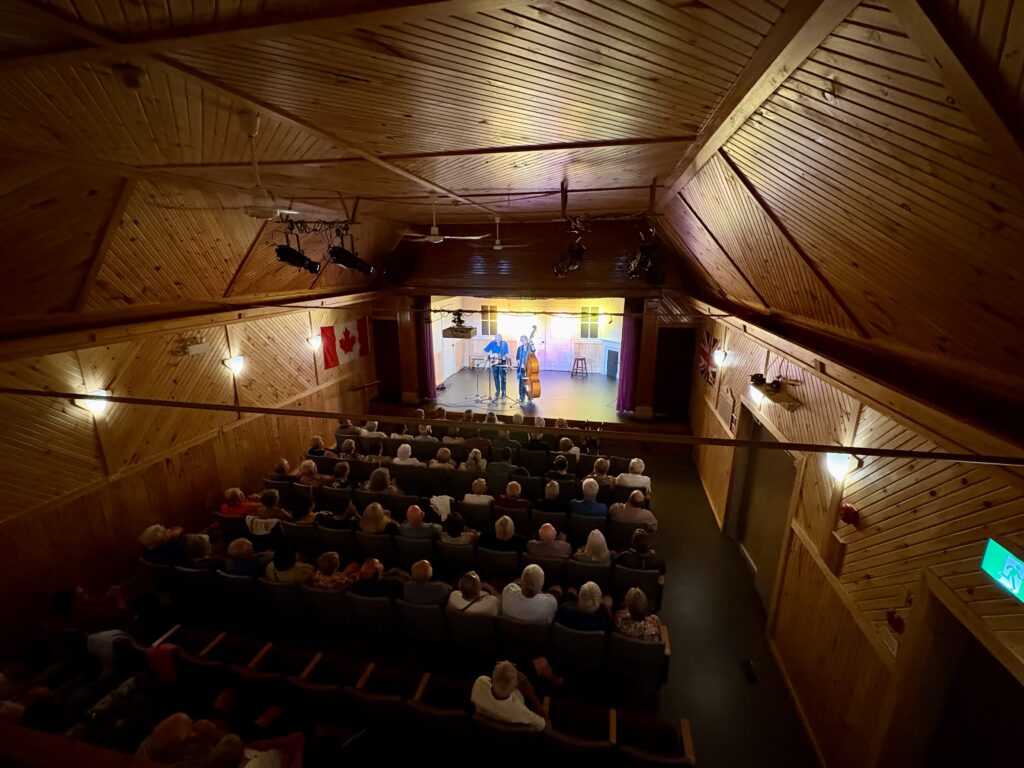 A pair of musicians on a theatre stage, with an audience watching them, surrounded by the wooden walls and ceiling of the theatre.