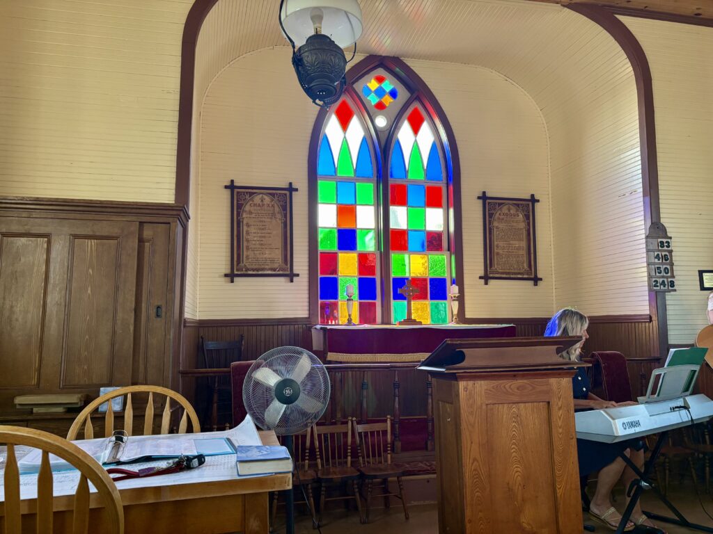 A colourful stained glass window in a church, the walls painted cream with wood trim, and a woman playing an electric piano on the right