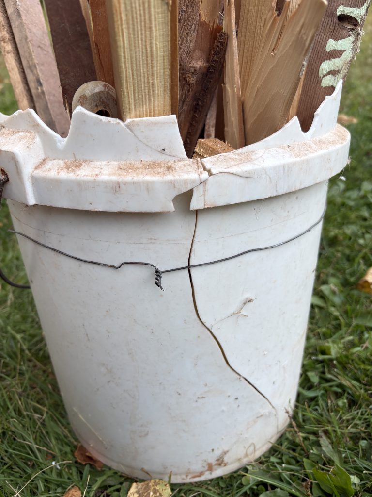 A cracked white plastic bucket with wood sticking out the top and a piece of wire wrapped around holding it together.
