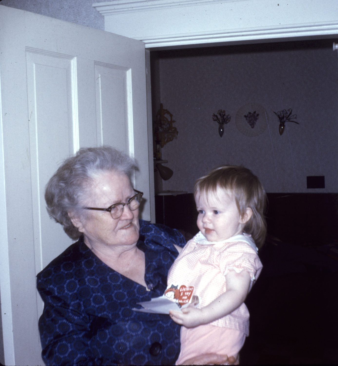 An older woman in a blue dress with grey hair and wearing glasses holding a small child in a pink outfit in front of a doorway