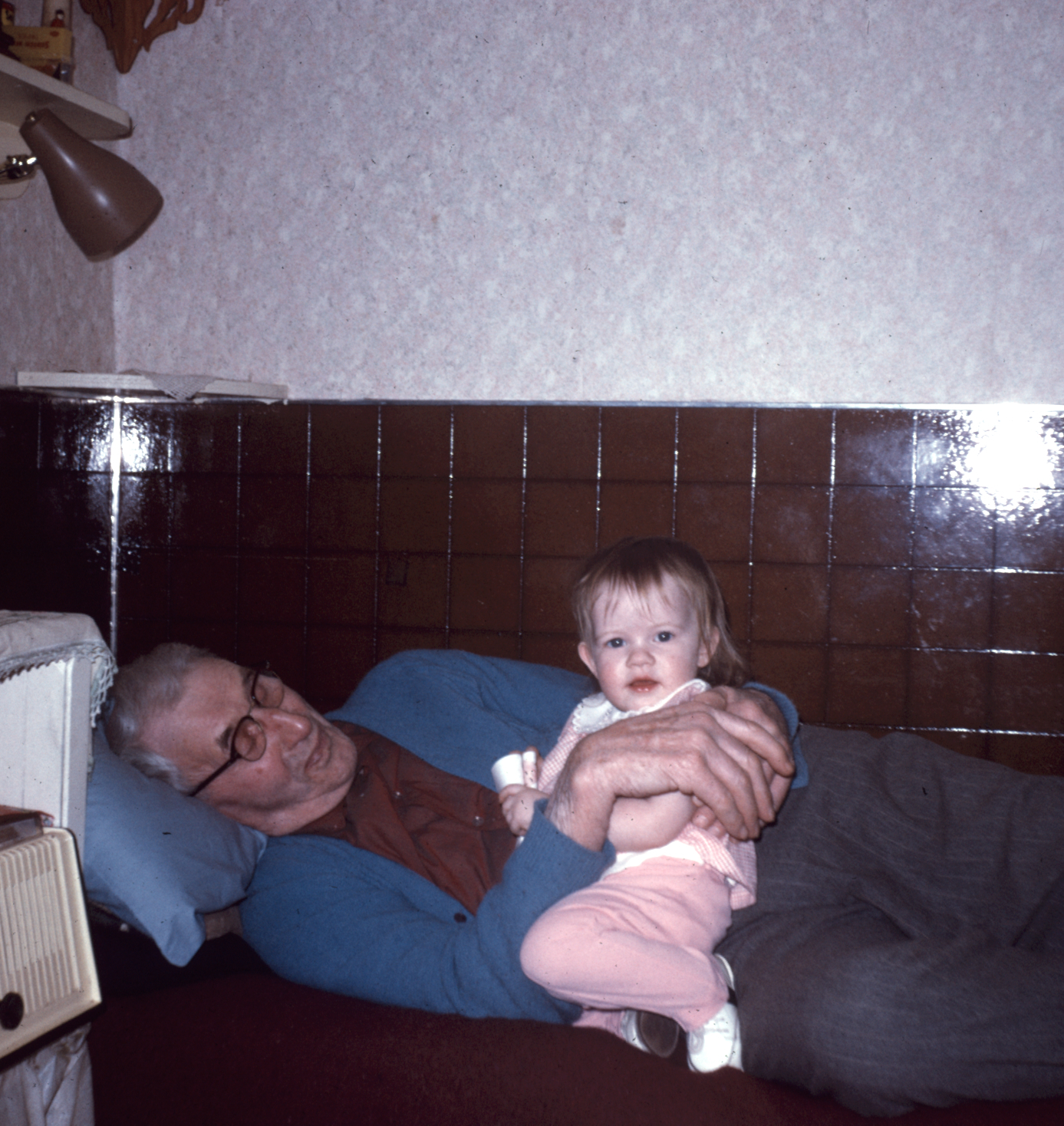 An older man reclining on a couch holding a small child in a pink outfit. A brown and white wall is behind them.