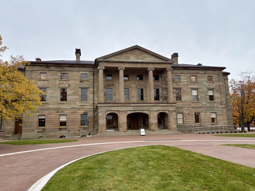 A large stone building with many windows and four columns, with a driveway and grass in the foreground.